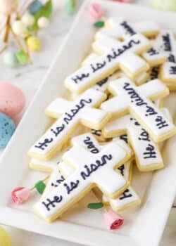 "He Is Risen" Cutout Cookies stacked on a white platter with decorative pink roses