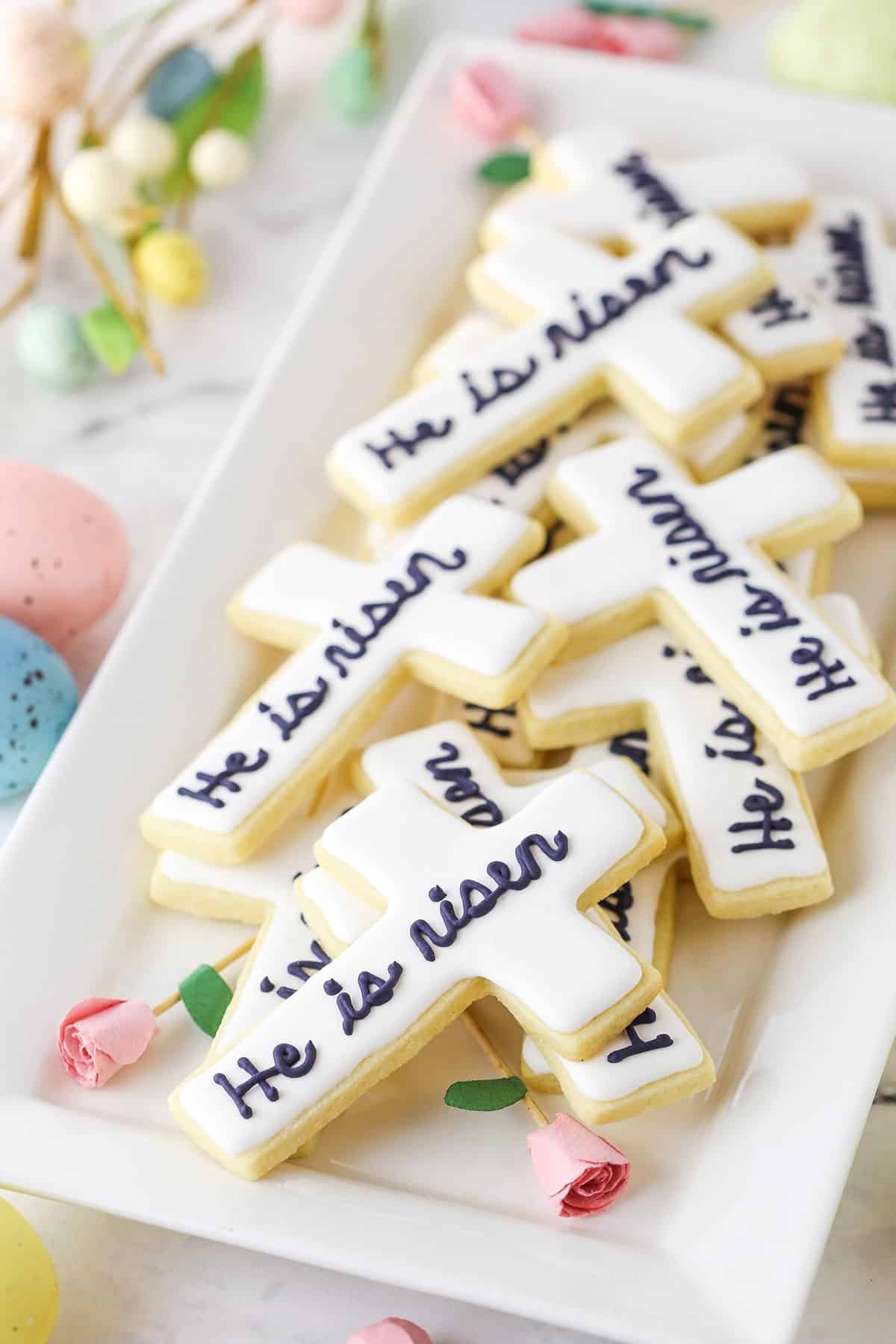 "He Is Risen" Cutout Cookies stacked on a white platter with decorative pink roses