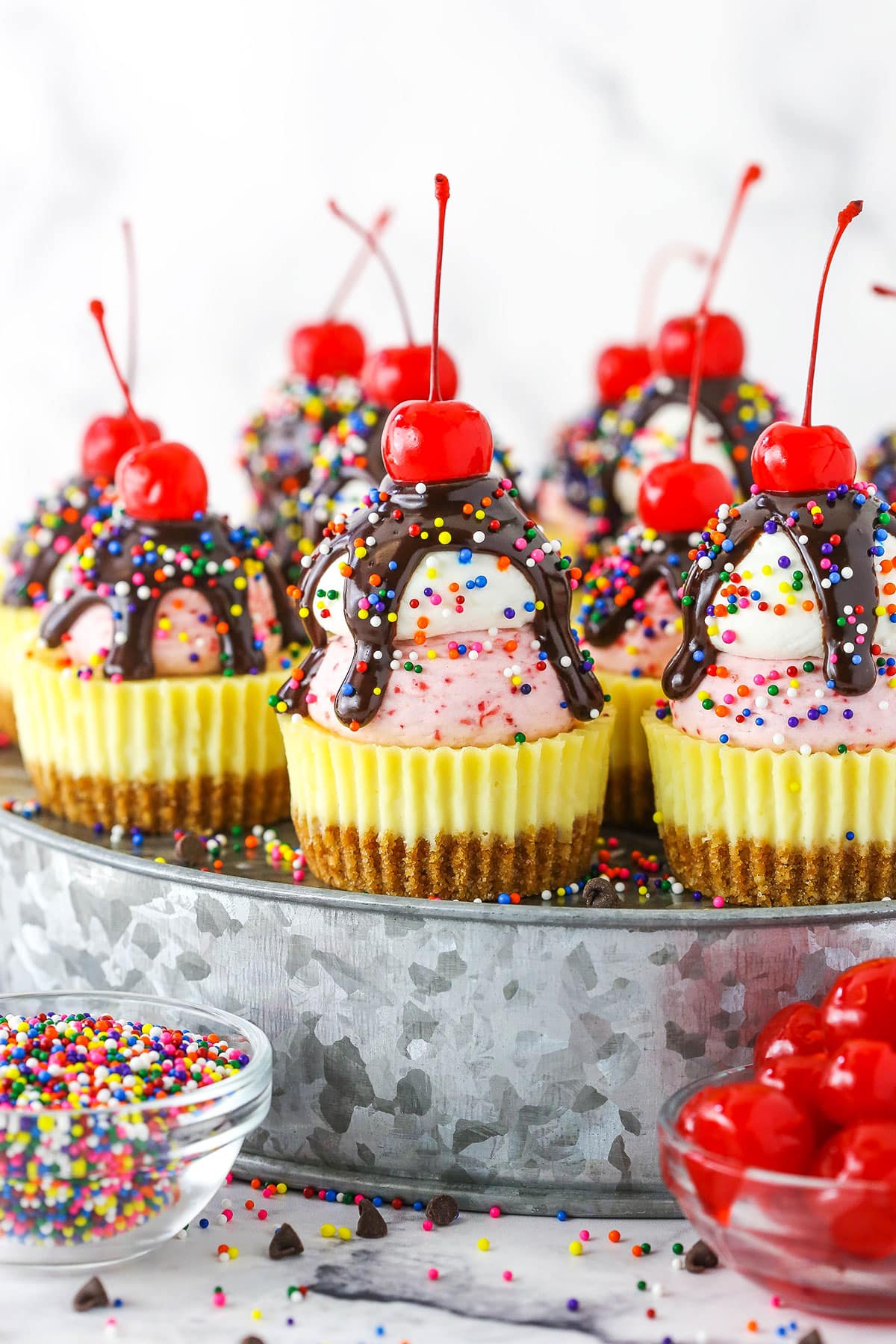 Ice Cream Sundae Mini Cheesecakes topped with chocolate drip and sprinkles with a cherry on top, on a metal platter