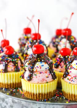 Ice Cream Sundae Mini Cheesecakes topped with chocolate drip and sprinkles with a cherry on top, on a metal platter