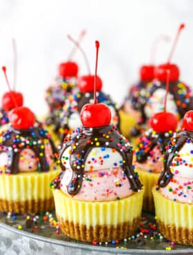 Ice Cream Sundae Mini Cheesecakes topped with chocolate drip and sprinkles with a cherry on top, on a metal platter