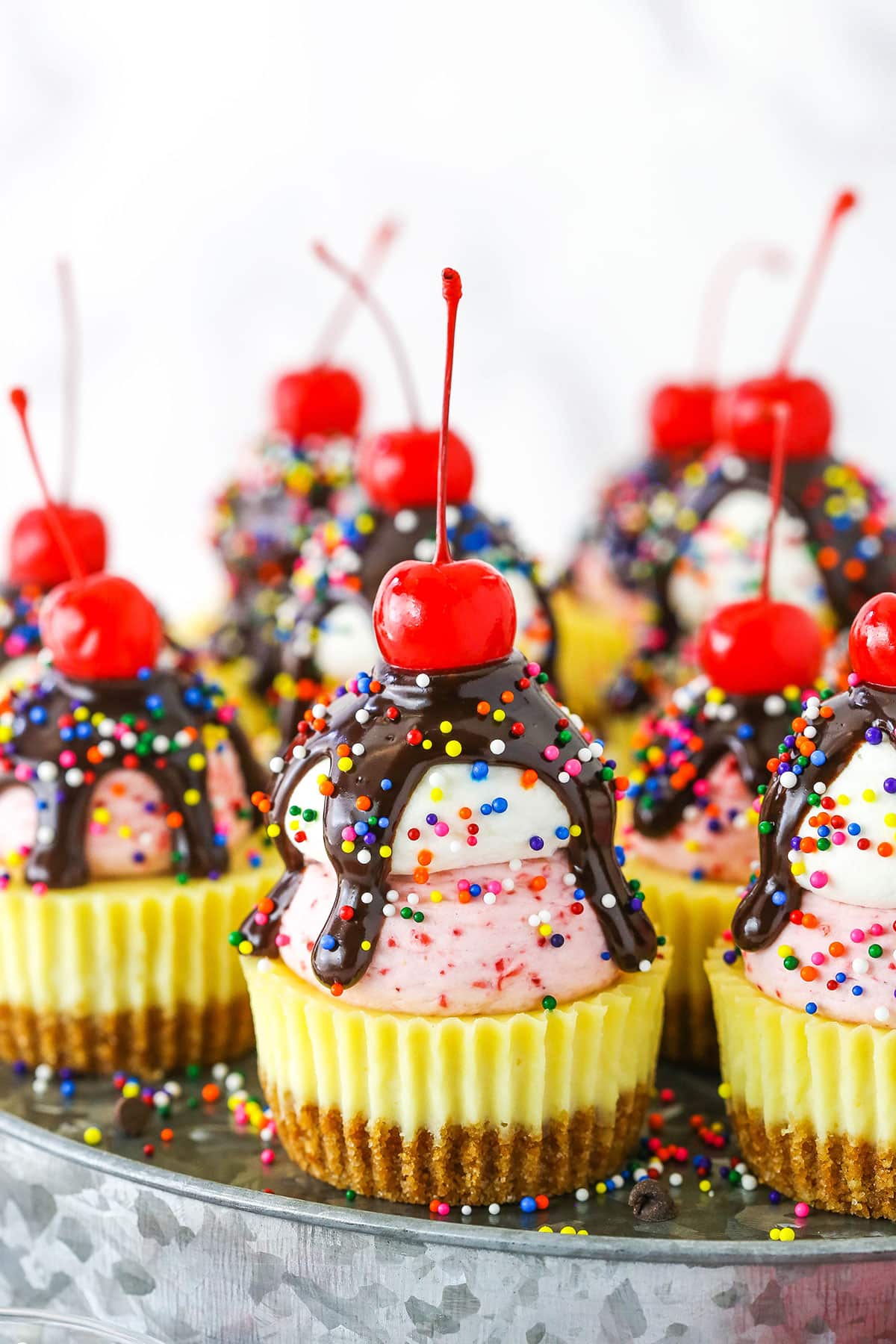 These Ice Cream Sundae Mini Cheesecakes are so fun and perfect for summer! The vanilla mini cheesecakes have an ice cream cone crust and are topped with strawberry and vanilla whipped cream, chocolate sauce, sprinkles and a cherry! They are pretty much guaranteed to bring a smile to your face! Ice Cream Sundae Mini Cheesecakes topped with chocolate drip and sprinkles with a cherry on top, on a metal platter