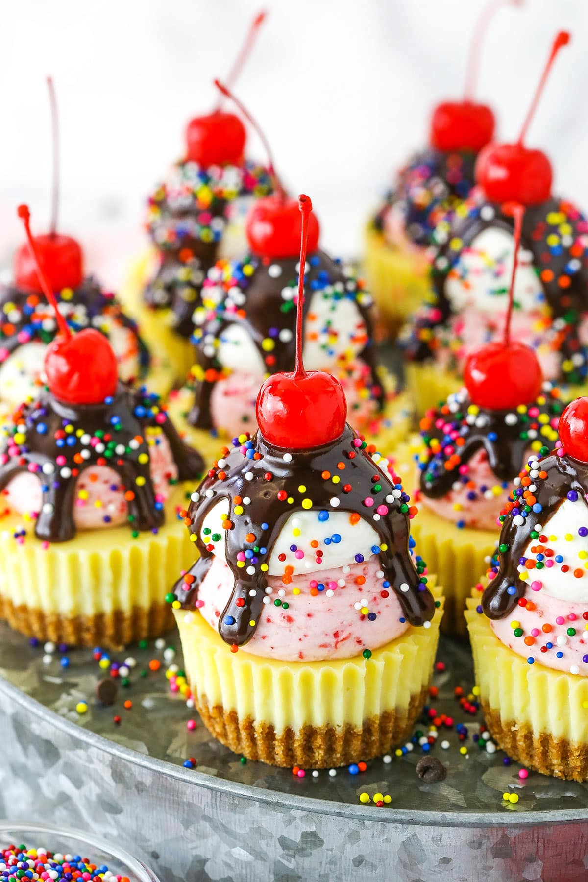 Ice Cream Sundae Mini Cheesecakes topped with chocolate drip and sprinkles with a cherry on top, on a metal platter