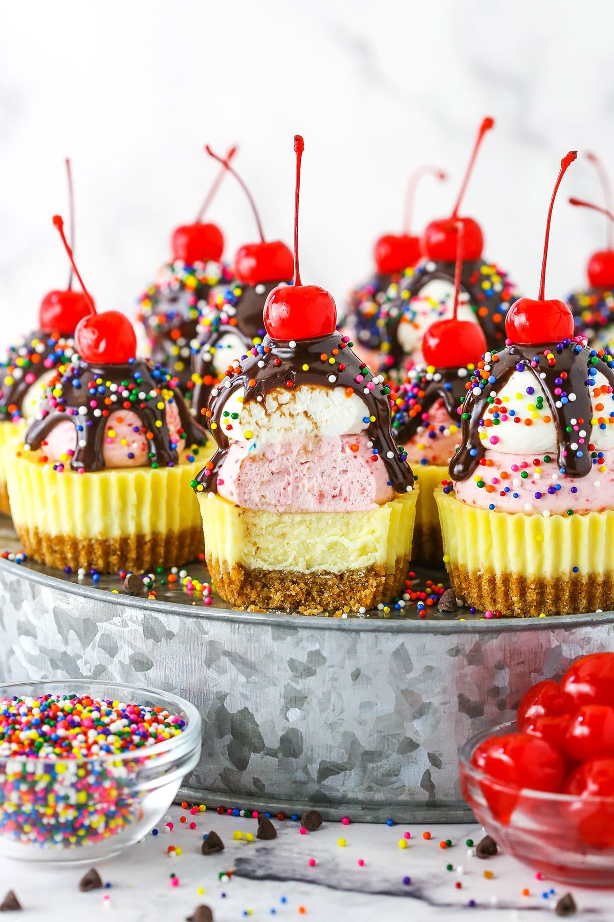 Ice Cream Sundae Mini Cheesecakes topped with chocolate drip and sprinkles with a cherry on top, on a metal platter