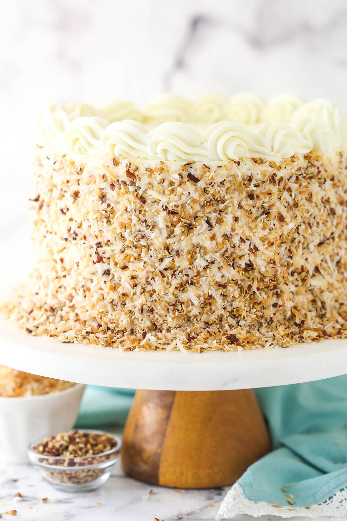 Italian cream cake on a cake stand near a bowl of toasted coconut and toasted pecans.