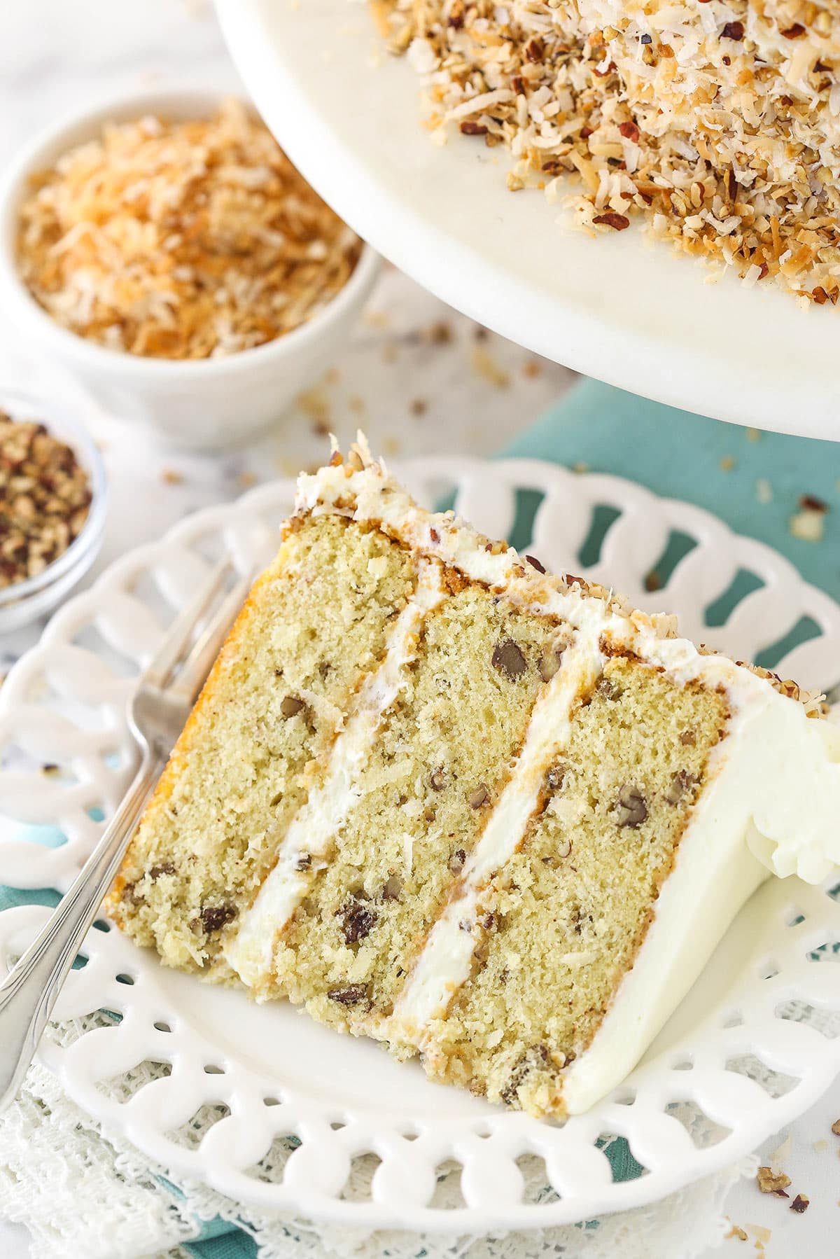 A slice of Italian cream cake on a plate with a fork near a bowl of toasted coconut and toasted pecans.