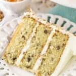 A slice of Italian cream cake on a plate with a fork near a bowl of toasted coconut and toasted pecans.