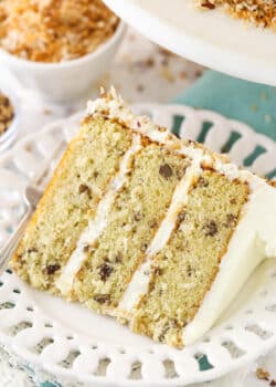 A slice of Italian cream cake on a plate with a fork near a bowl of toasted coconut and toasted pecans.