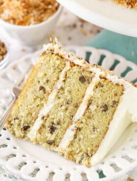 A slice of Italian cream cake on a plate with a fork near a bowl of toasted coconut and toasted pecans.