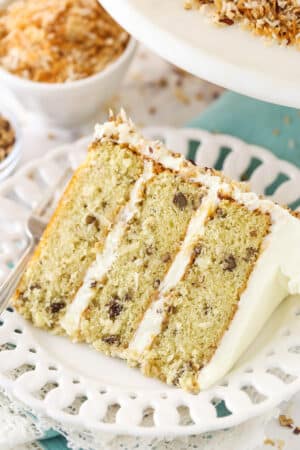 A slice of Italian cream cake on a plate with a fork near a bowl of toasted coconut and toasted pecans.