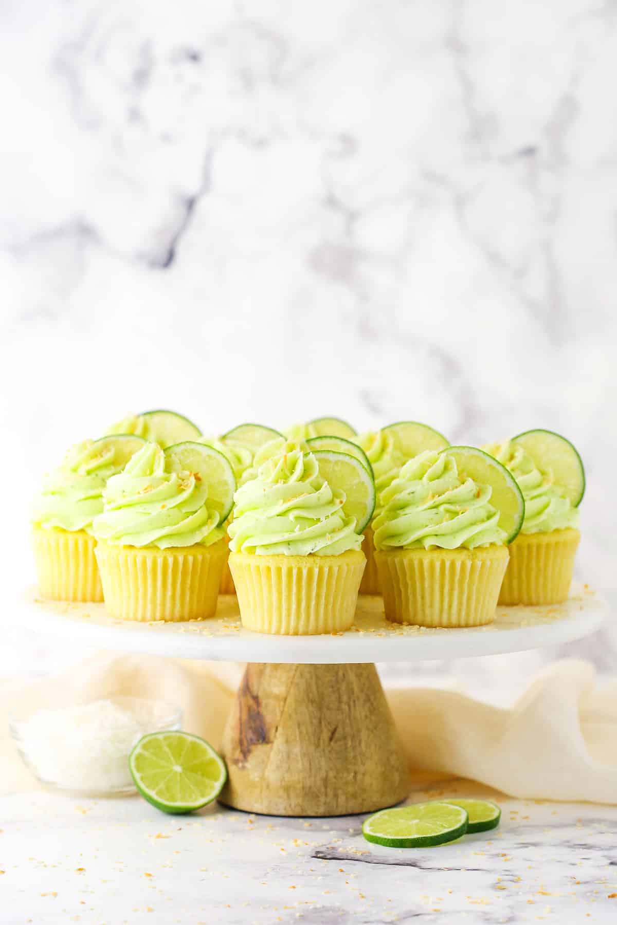 These key lime coconut cupcakes are made of soft, fluffy coconut cupcakes topped with zesty key lime buttercream. They're a tropical dream! Key lime coconut cupcakes on a cake stand with key lime slices underneath.
