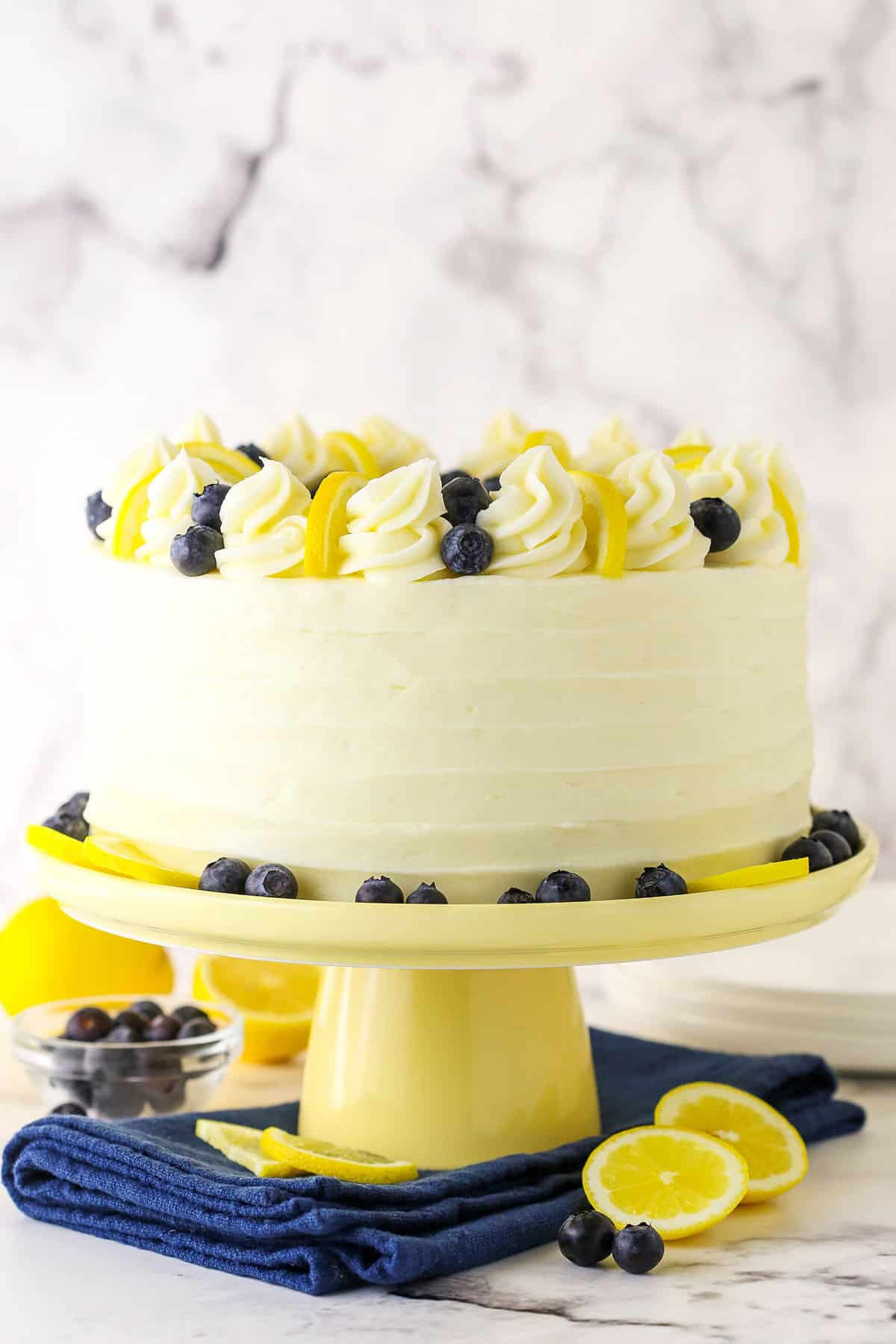 Side view of a full Lemon Blueberry Layer Cake with white swirls, blueberries and cut lemons on a yellow cake stand