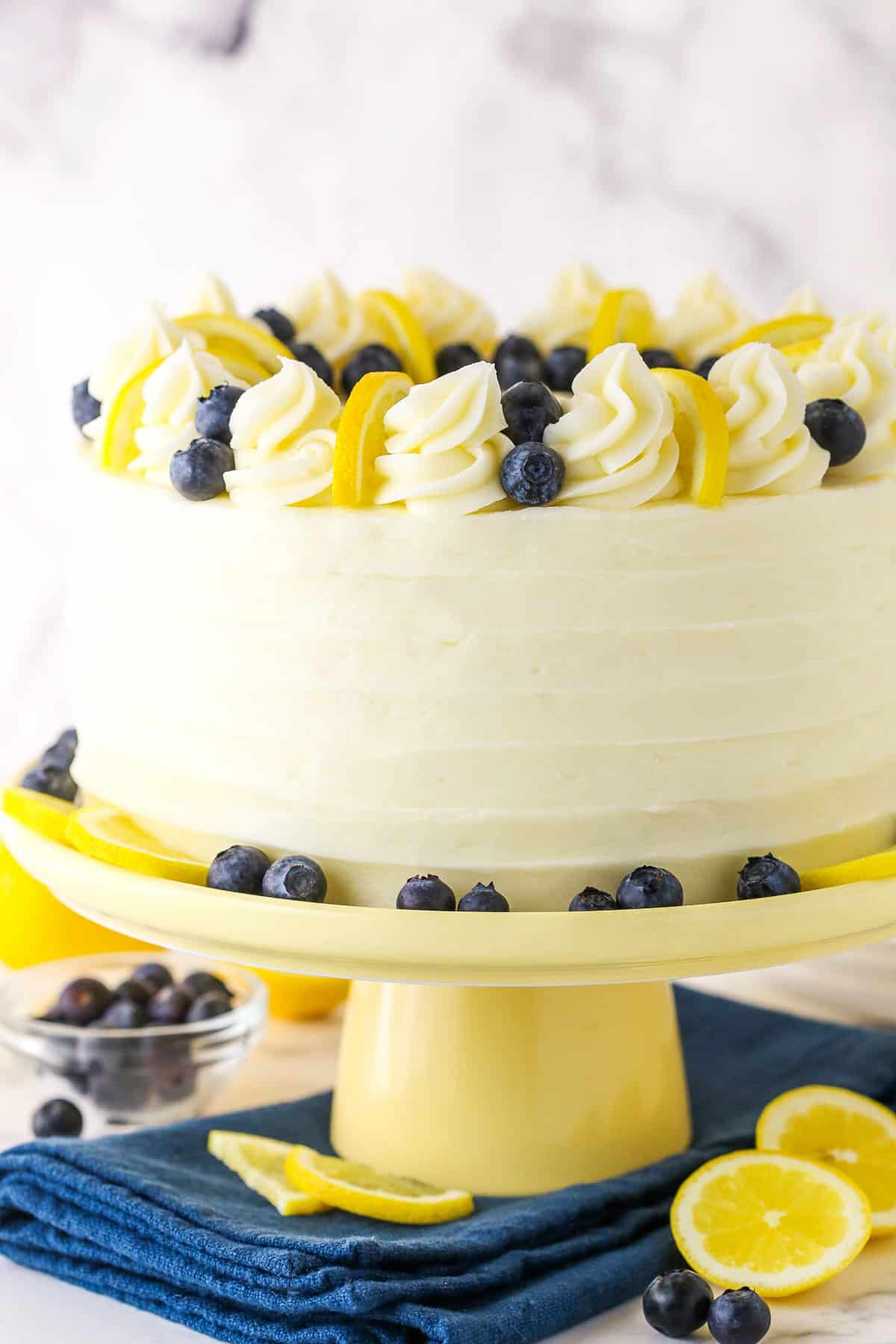 Side view of a full Lemon Blueberry Layer Cake with white swirls, blueberries and cut lemons on a yellow cake stand