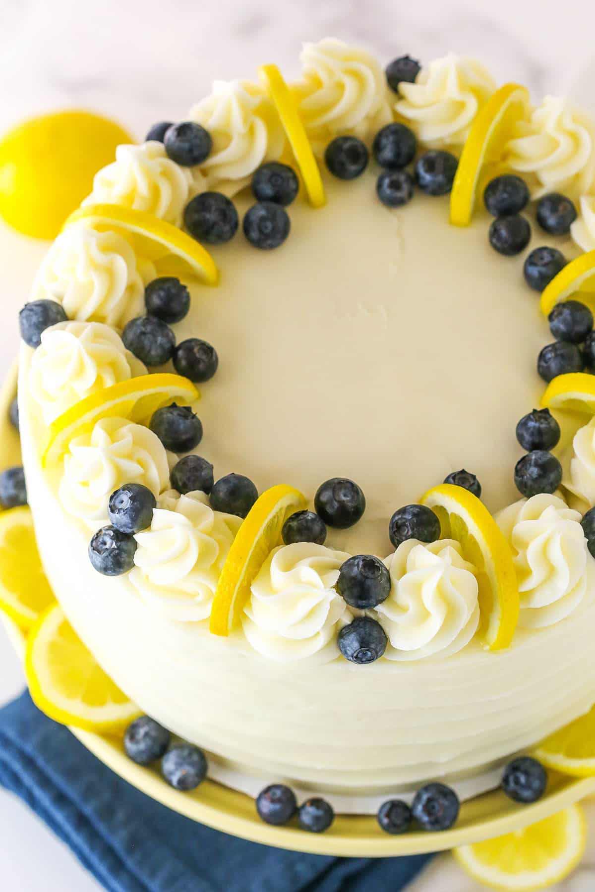 Overhead view of a full Lemon Blueberry Layer Cake with white swirls, blueberries and cut lemons on a yellow cake stand
