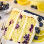 A slice of Lemon Blueberry Layer Cake with a fork on a white plate and blueberries and cut lemons in the background