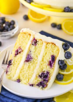 A slice of Lemon Blueberry Layer Cake with a fork on a white plate and blueberries and cut lemons in the background