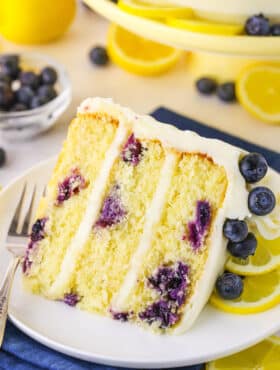 A slice of Lemon Blueberry Layer Cake with a fork on a white plate and blueberries and cut lemons in the background