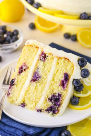 A slice of Lemon Blueberry Layer Cake with a fork on a white plate and blueberries and cut lemons in the background