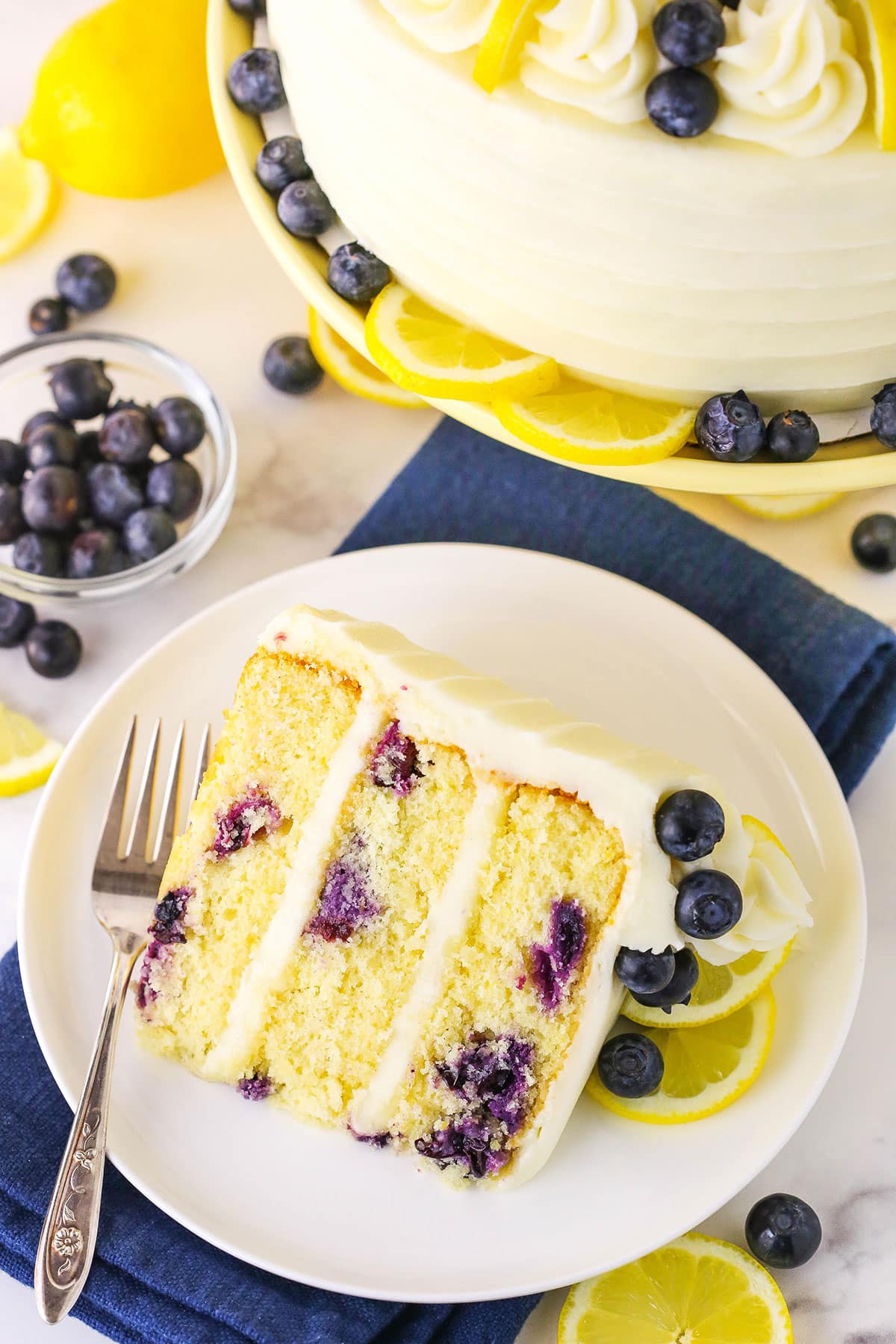 A slice of Lemon Blueberry Layer Cake with a fork on a white plate and blueberries and cut lemons in the background