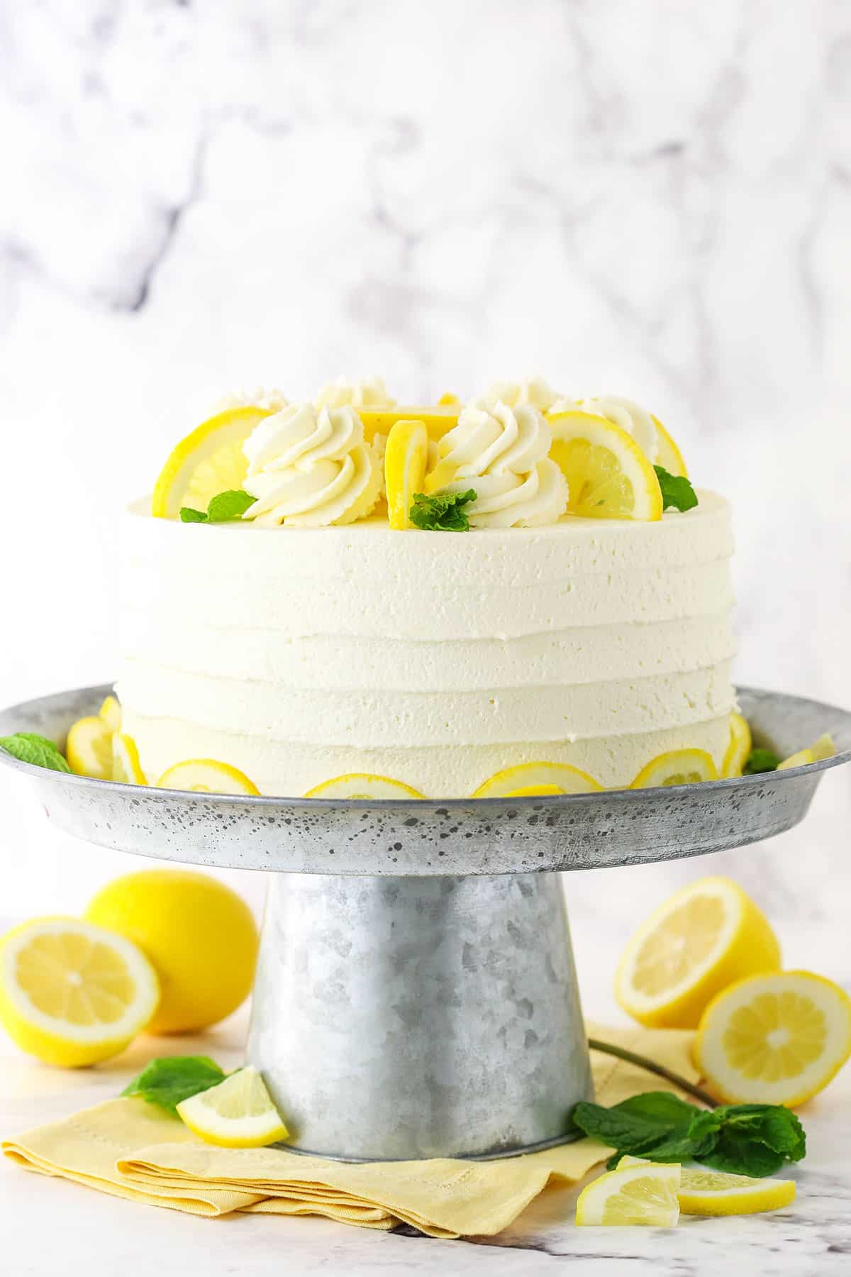 Side view of a full Lemon Mascarpone Layer Cake topped with white swirls and cut lemons on a metal colored cake stand