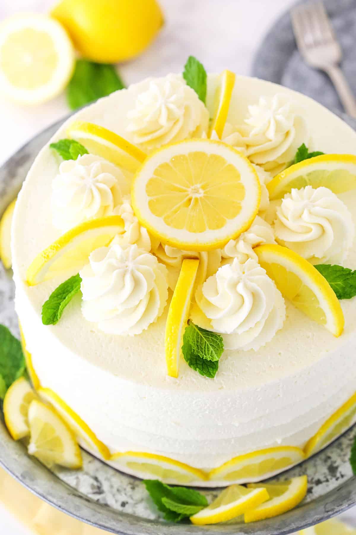 This Lemon Mascarpone Layer Cake is made with a moist lemon cake, lemon curd and a smooth and creamy whipped mascarpone frosting! It's light, yet sweet and tart and so darn good! It'll be your new favorite cake for spring! Overhead view of a full Lemon Mascarpone Layer Cake topped with white swirls and cut lemons on a metal colored cake stand