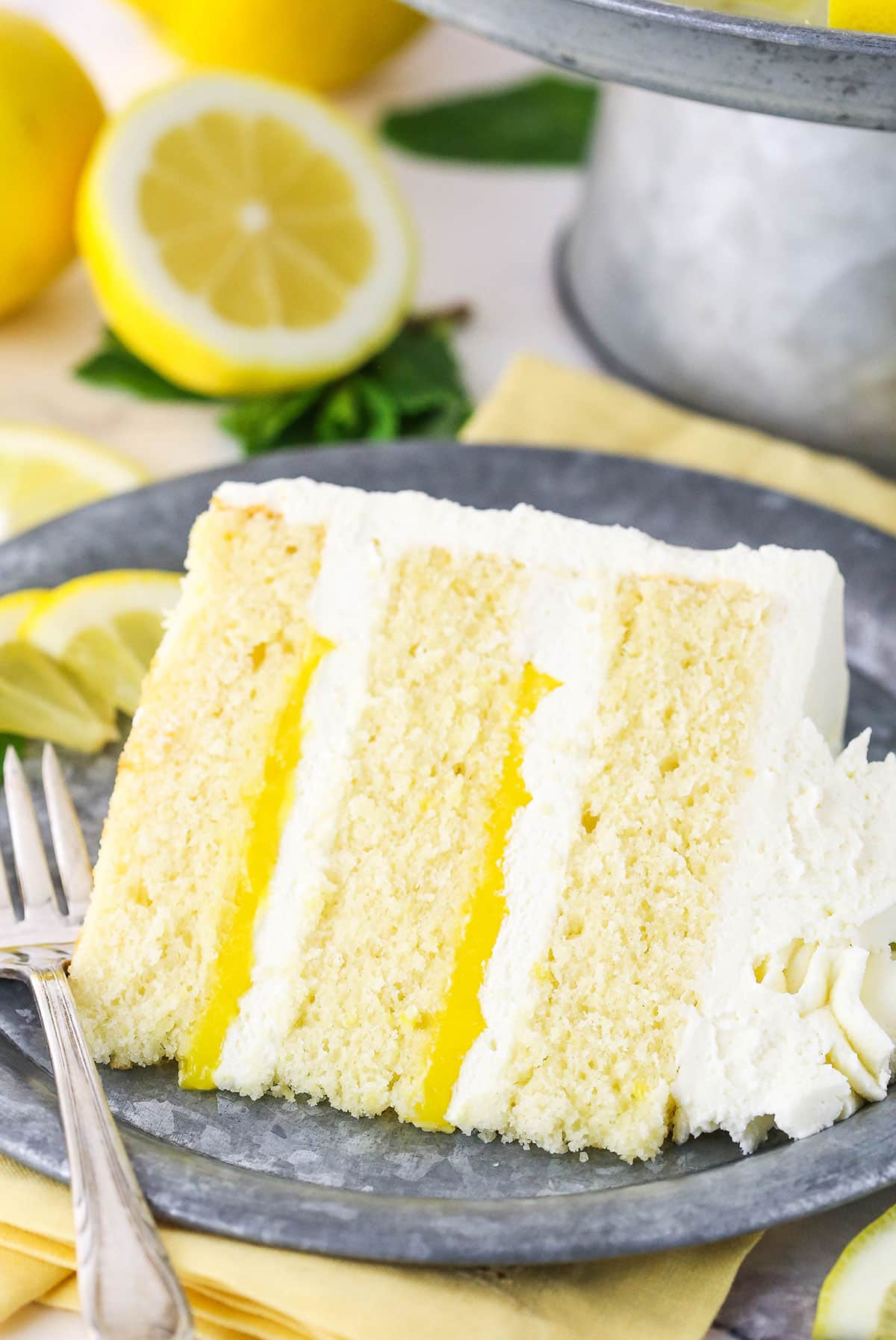 A slice of Lemon Mascarpone Layer Cake on a metal colored plate with a fork and cut lemons in the background
