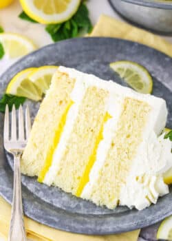 A slice of Lemon Mascarpone Layer Cake on a metal colored plate with a fork and cut lemons in the background