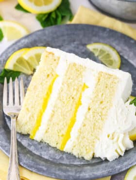 A slice of Lemon Mascarpone Layer Cake on a metal colored plate with a fork and cut lemons in the background