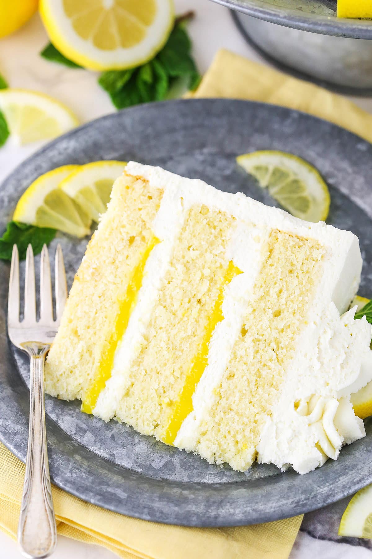 A slice of Lemon Mascarpone Layer Cake on a metal colored plate with a fork and cut lemons in the background