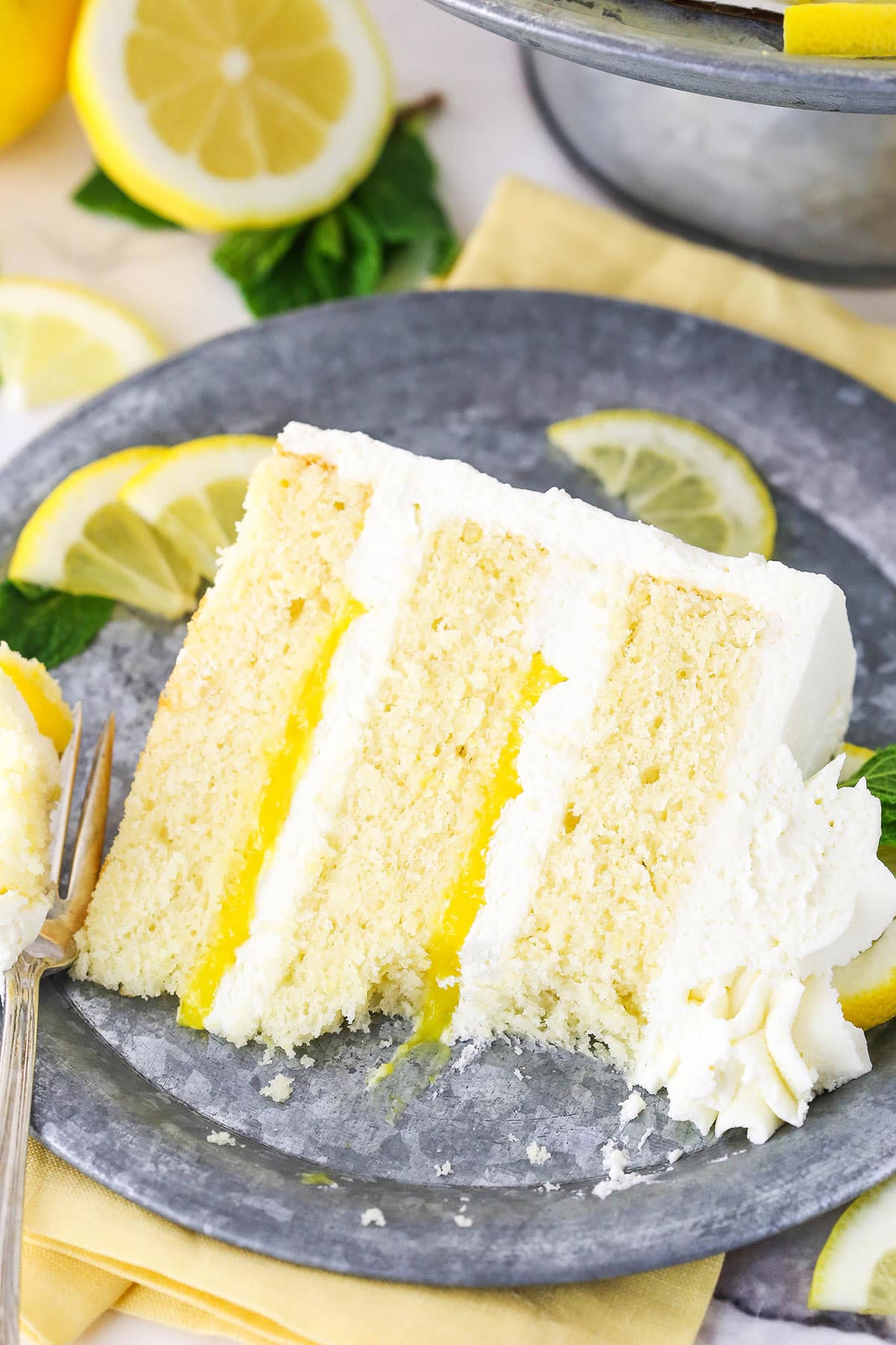 A slice of Lemon Mascarpone Layer Cake with a bite taken out on a metal colored plate with a fork and cut lemons in the background