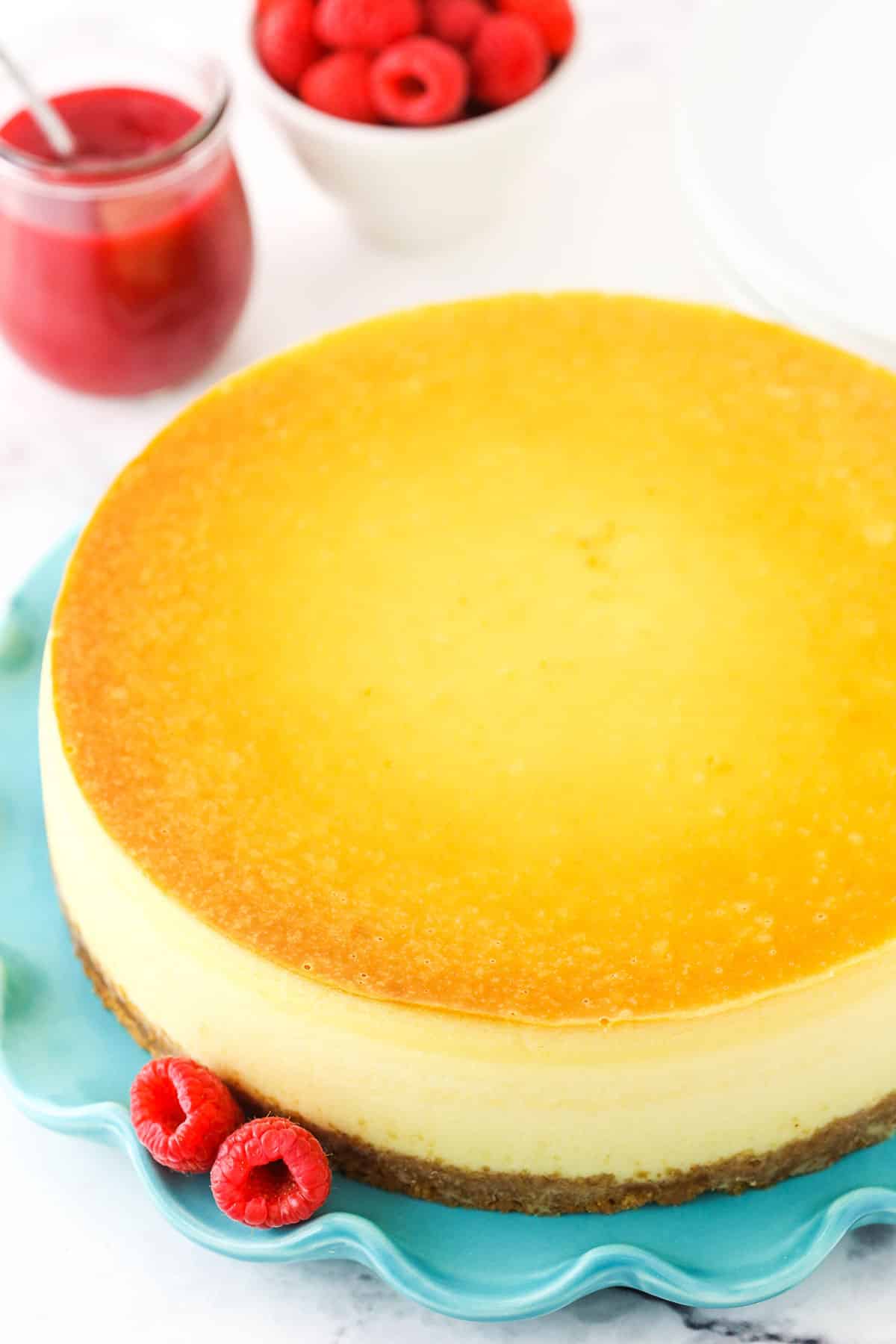 Overhead of full New York cheesecake on a blue platter with raspberries and raspberry sauce in the background.