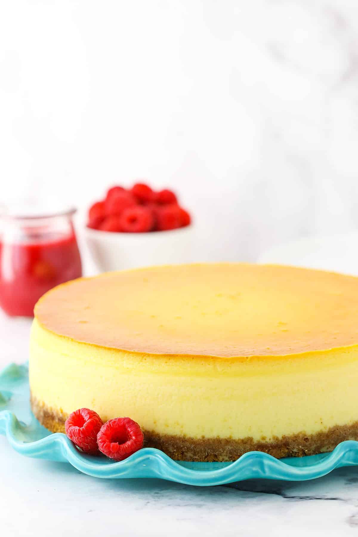 Full New York Cheesecake on a blue platter with raspberries and raspberry sauce in the background.