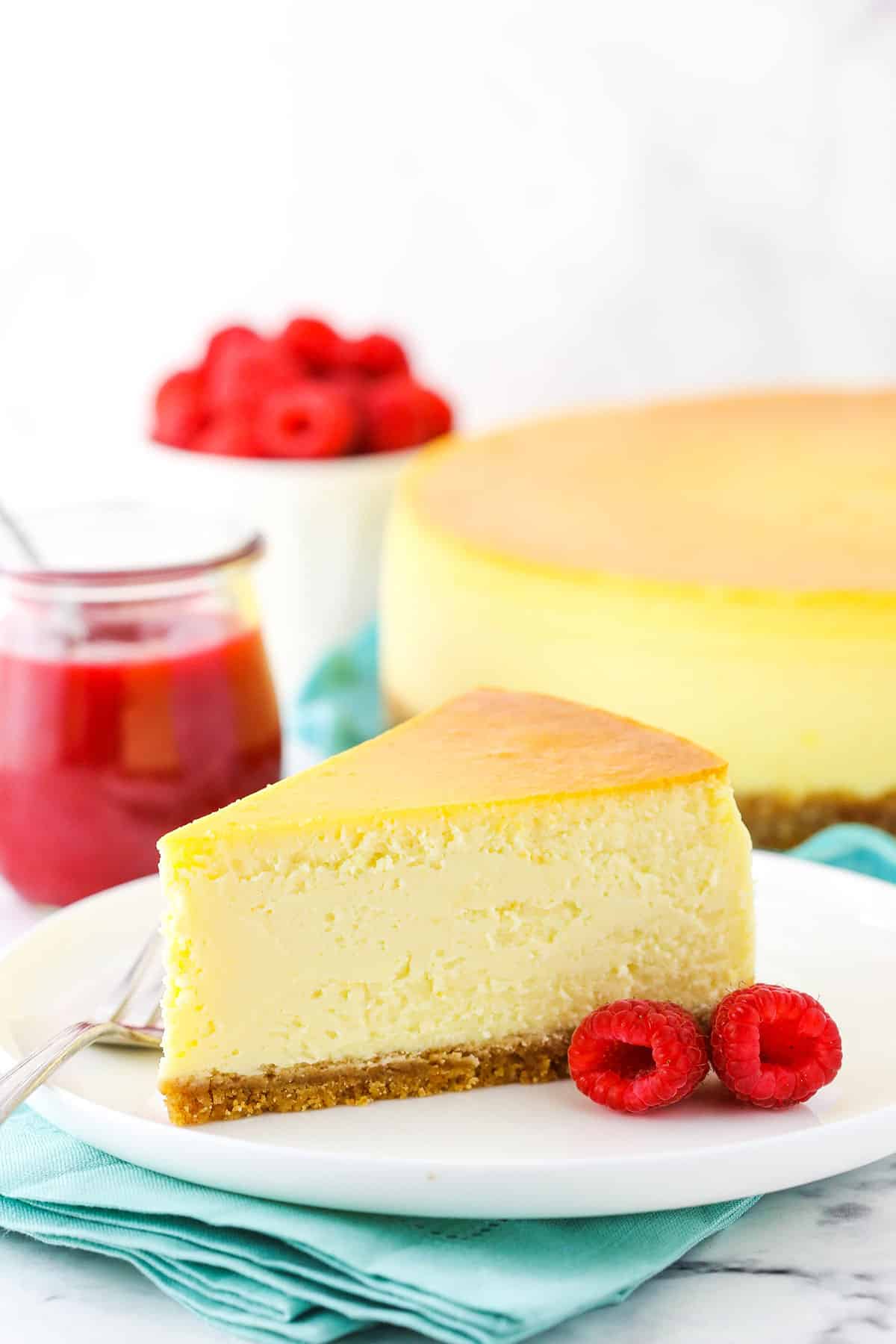 Slice of New York Style Cheesecake on a white plate with a fork and raspberries.