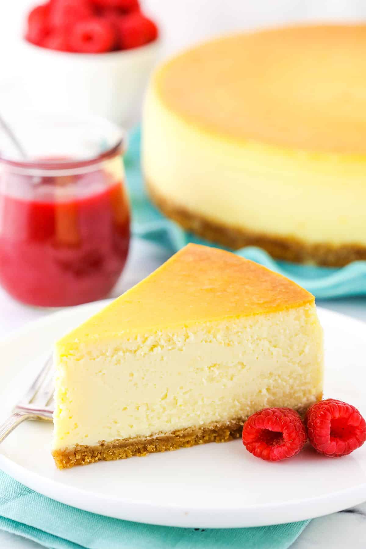 Slice of New York Style Cheesecake on a white plate with a fork and raspberries.