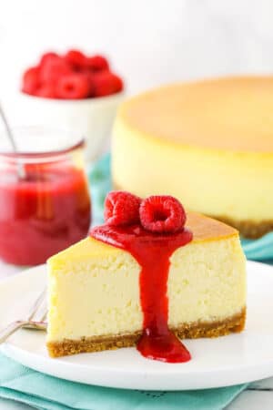 Slice of New York Style Cheesecake topped with raspberry sauce and raspberries on a white plate with a fork.