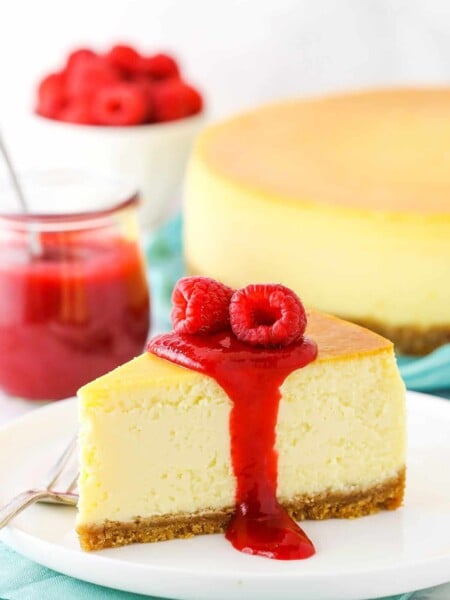 Slice of New York Style Cheesecake topped with raspberry sauce and raspberries on a white plate with a fork.