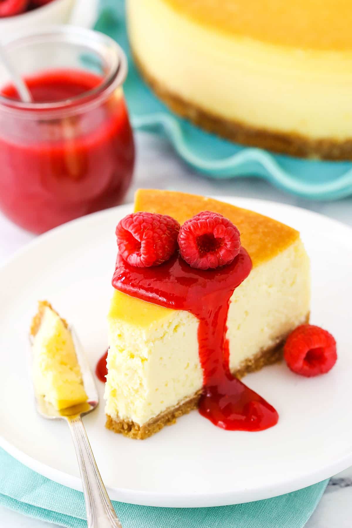 Slice of New York Style Cheesecake topped with raspberry sauce and raspberries on a white plate with a fork.