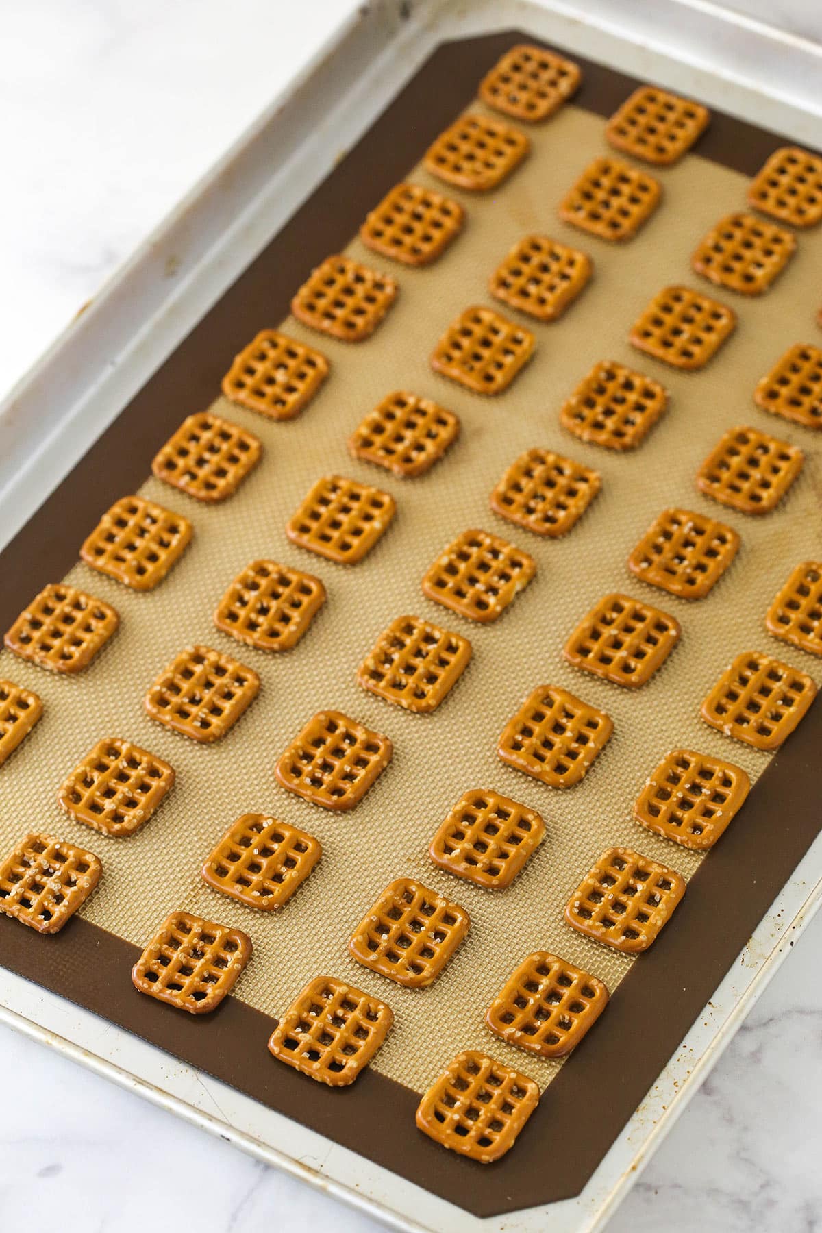 These Pretzel M&M Hugs are festive little chocolate treats! They are easy to make, use only 3 ingredients and you can easily change up the colors for any holiday! lots of square pretzels laid out on silicone baking mat on baking sheet