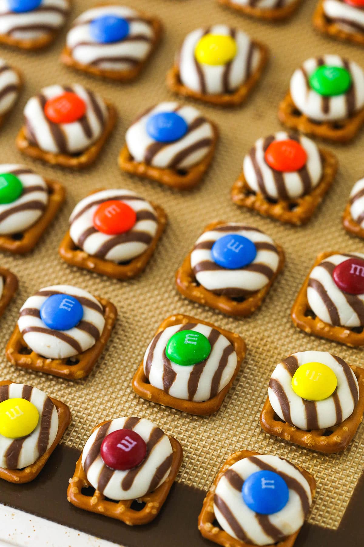 These Pretzel M&M Hugs are festive little chocolate treats! They are easy to make, use only 3 ingredients and you can easily change up the colors for any holiday! finished pretzel hugs with M&MS on top on silicone baking mat