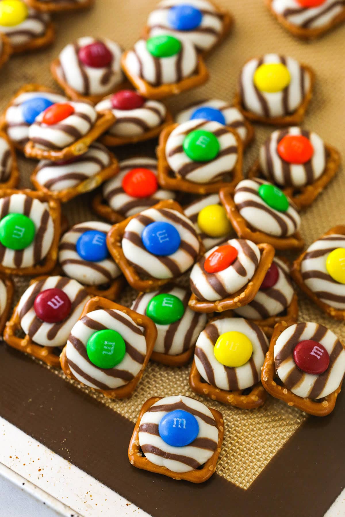 These Pretzel M&M Hugs are festive little chocolate treats! They are easy to make, use only 3 ingredients and you can easily change up the colors for any holiday! a pile of pretzel M&M hugs on a silicon baking mat lined baking sheet