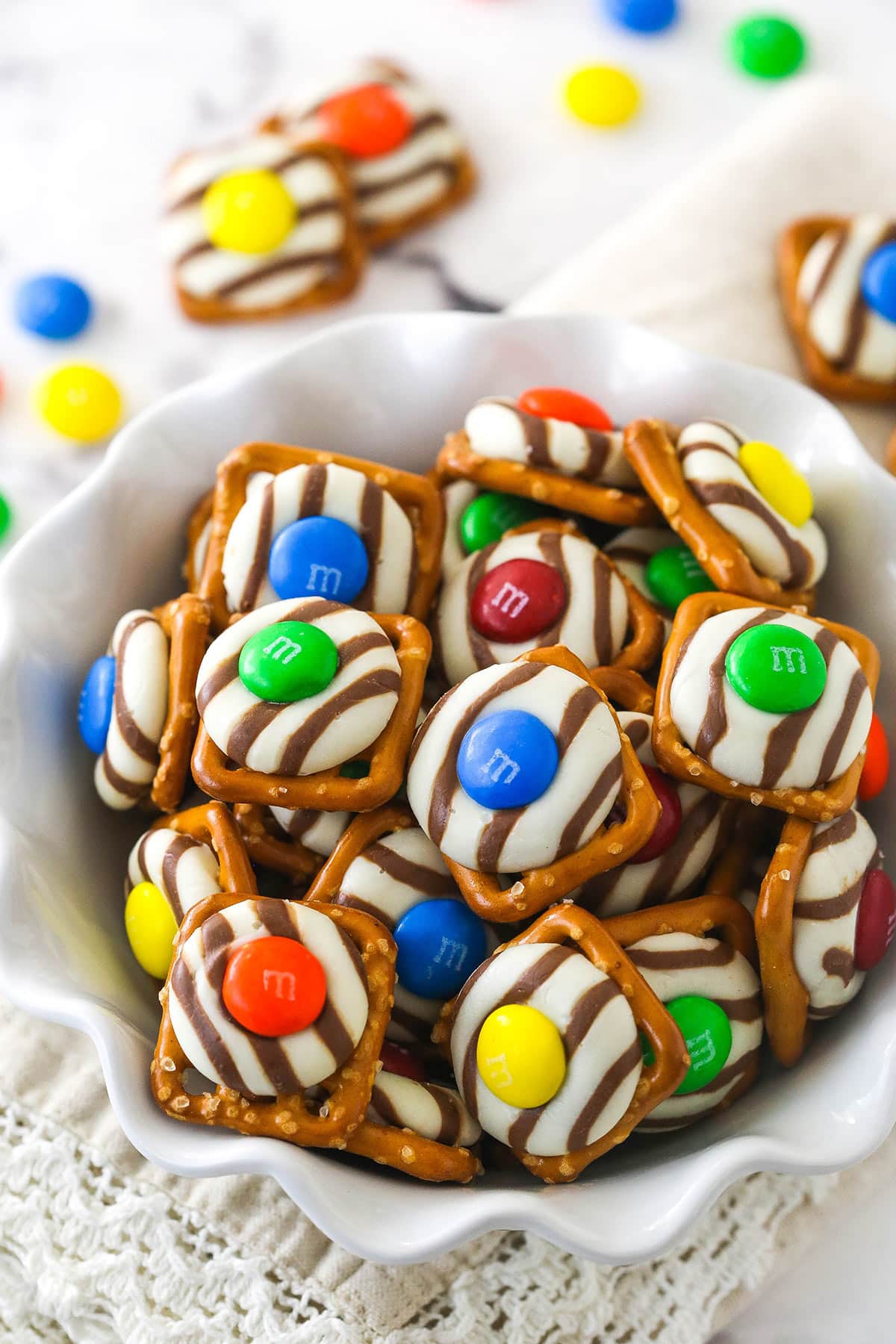 These Pretzel M&M Hugs are festive little chocolate treats! They are easy to make, use only 3 ingredients and you can easily change up the colors for any holiday! colorful pretzel hugs in a white ruffle bowl