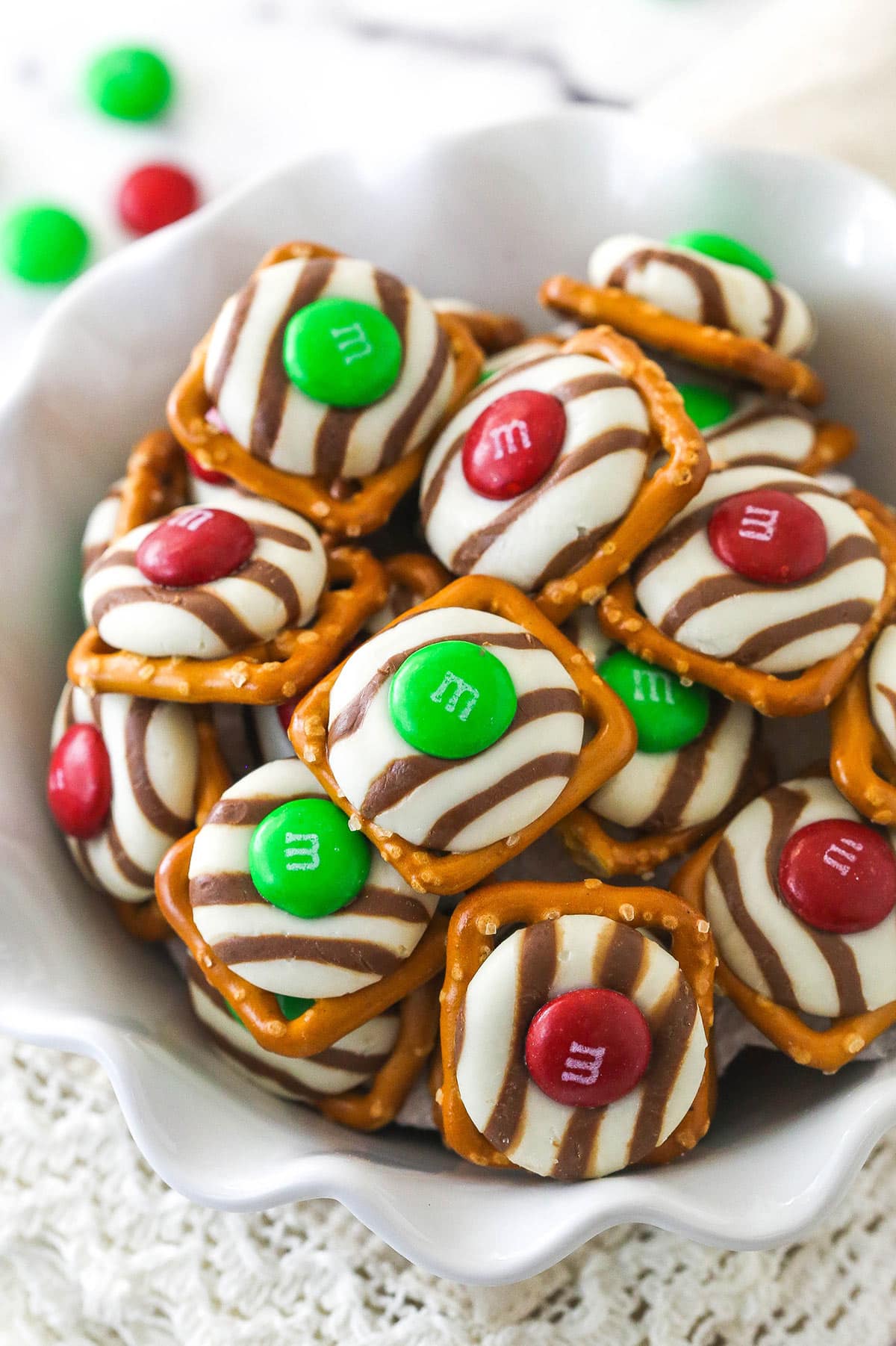 These Pretzel M&M Hugs are festive little chocolate treats! They are easy to make, use only 3 ingredients and you can easily change up the colors for any holiday! red and green Christmas pretzel M&M hugs in a white ruffle bowl
