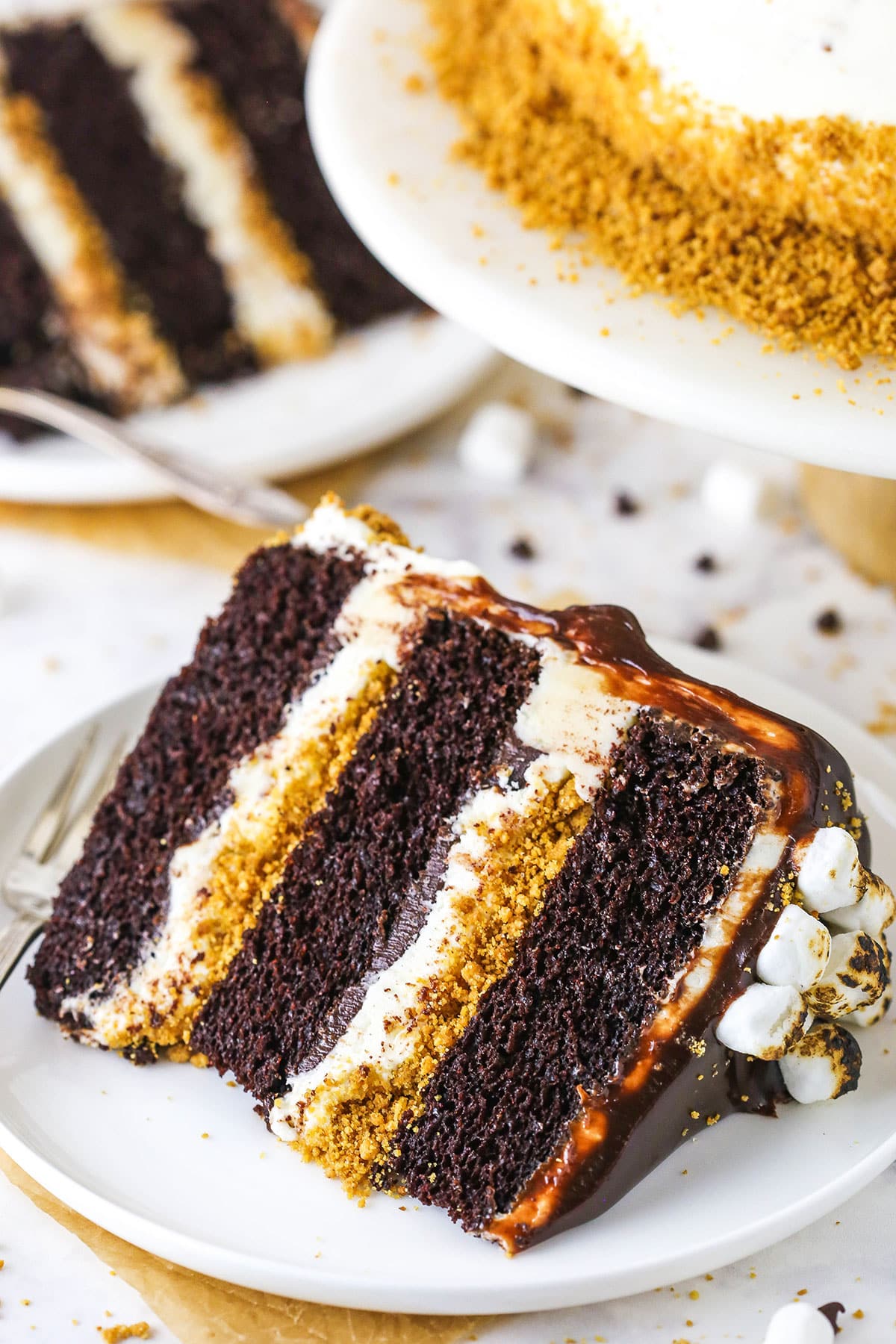 This S’mores Cake is an amazing combination of moist chocolate cake, toasted marshmallow buttercream, graham cracker crunchies and chocolate ganache! A slice of S'mores Cake with a fork on a white plate