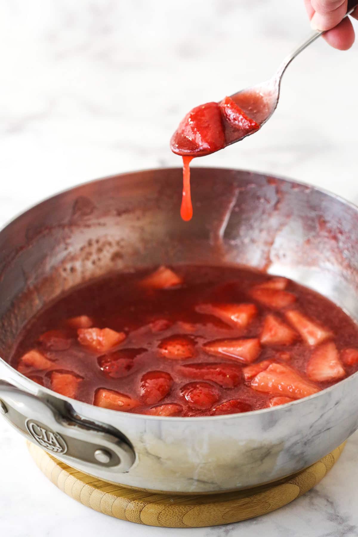This perfect-for-summer strawberry cheesecake is made of a Graham Cracker crust, a creamy cheesecake filling, and a sweet strawberry sauce. Strawberry sauce in a saucepan with a spoon dipping into it.