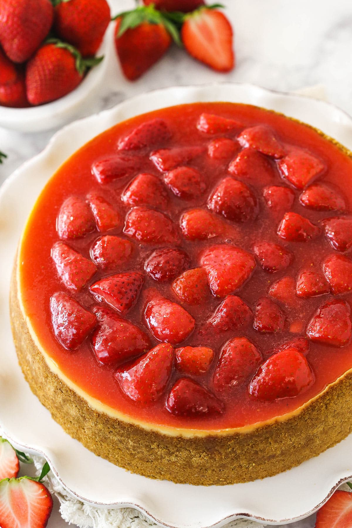 This perfect-for-summer strawberry cheesecake is made of a Graham Cracker crust, a creamy cheesecake filling, and a sweet strawberry sauce. Overhead of strawberry cheesecake on a serving platter with fresh strawberries scattered around.