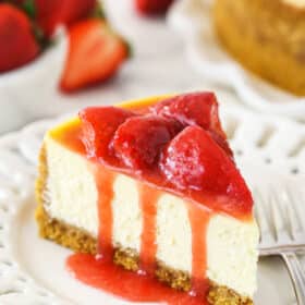 As slice of ultimate strawberry cheesecake on a plate near a bowl of fresh strawberries and a full strawberry cheesecake.