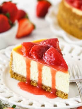 As slice of ultimate strawberry cheesecake on a plate near a bowl of fresh strawberries and a full strawberry cheesecake.