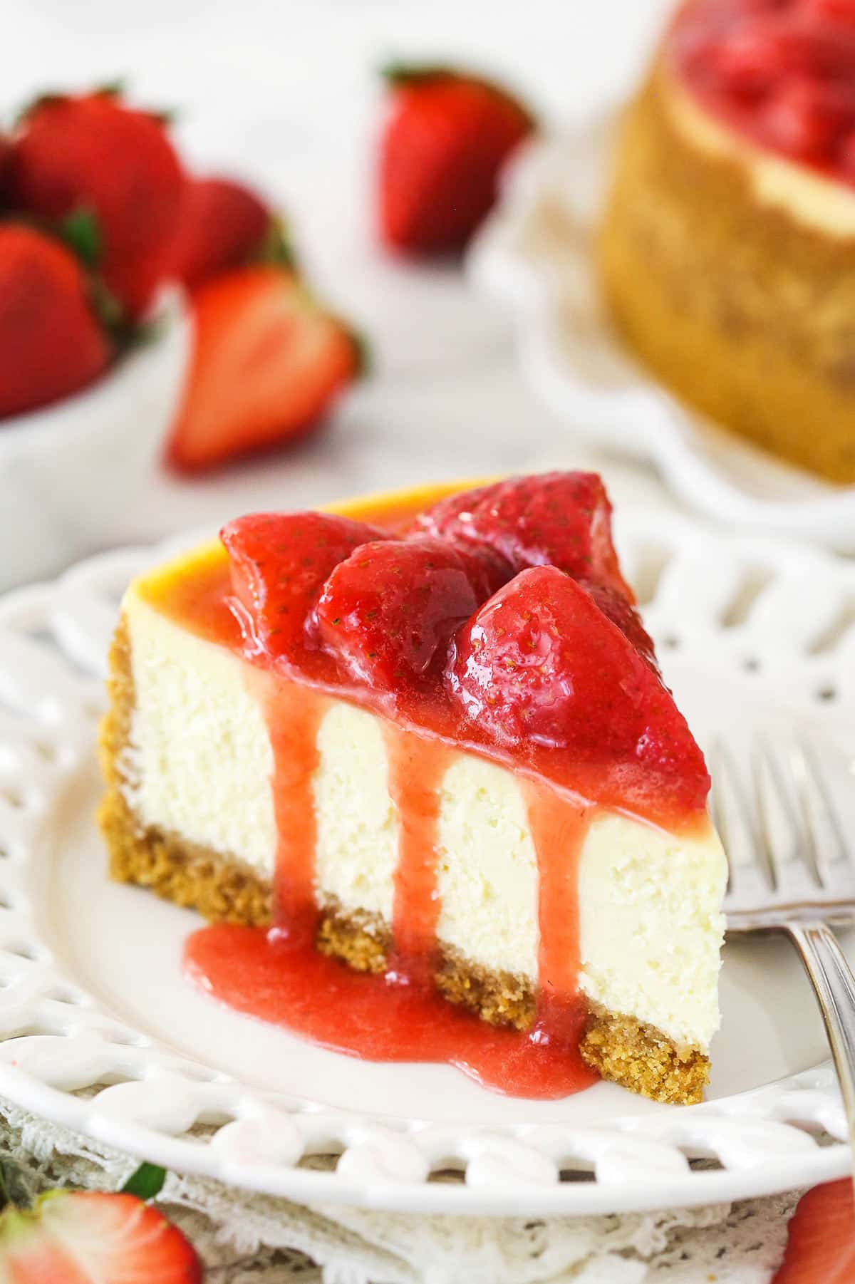 As slice of ultimate strawberry cheesecake on a plate near a bowl of fresh strawberries and a full strawberry cheesecake.