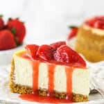 A slice of strawberry cheesecake on a plate with a fork near fresh strawberries and a full strawberry cheesecake.