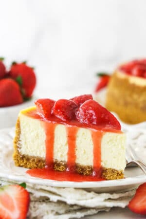 A slice of strawberry cheesecake on a plate with a fork near fresh strawberries and a full strawberry cheesecake.